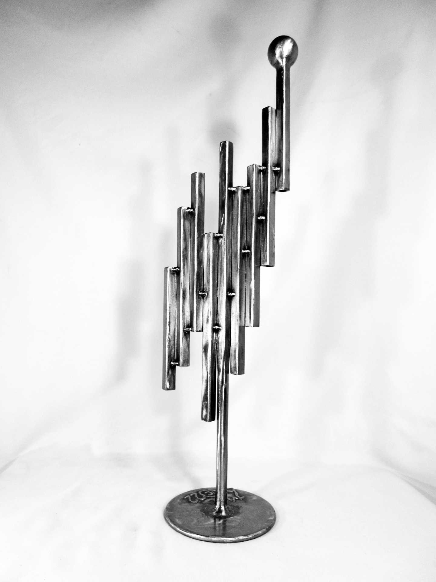 Metal sculpture featuring a vertical arrangement of staggered rectangular elements rising upward along a central rod, topped with a sphere on a circular base. By Michael Herold.