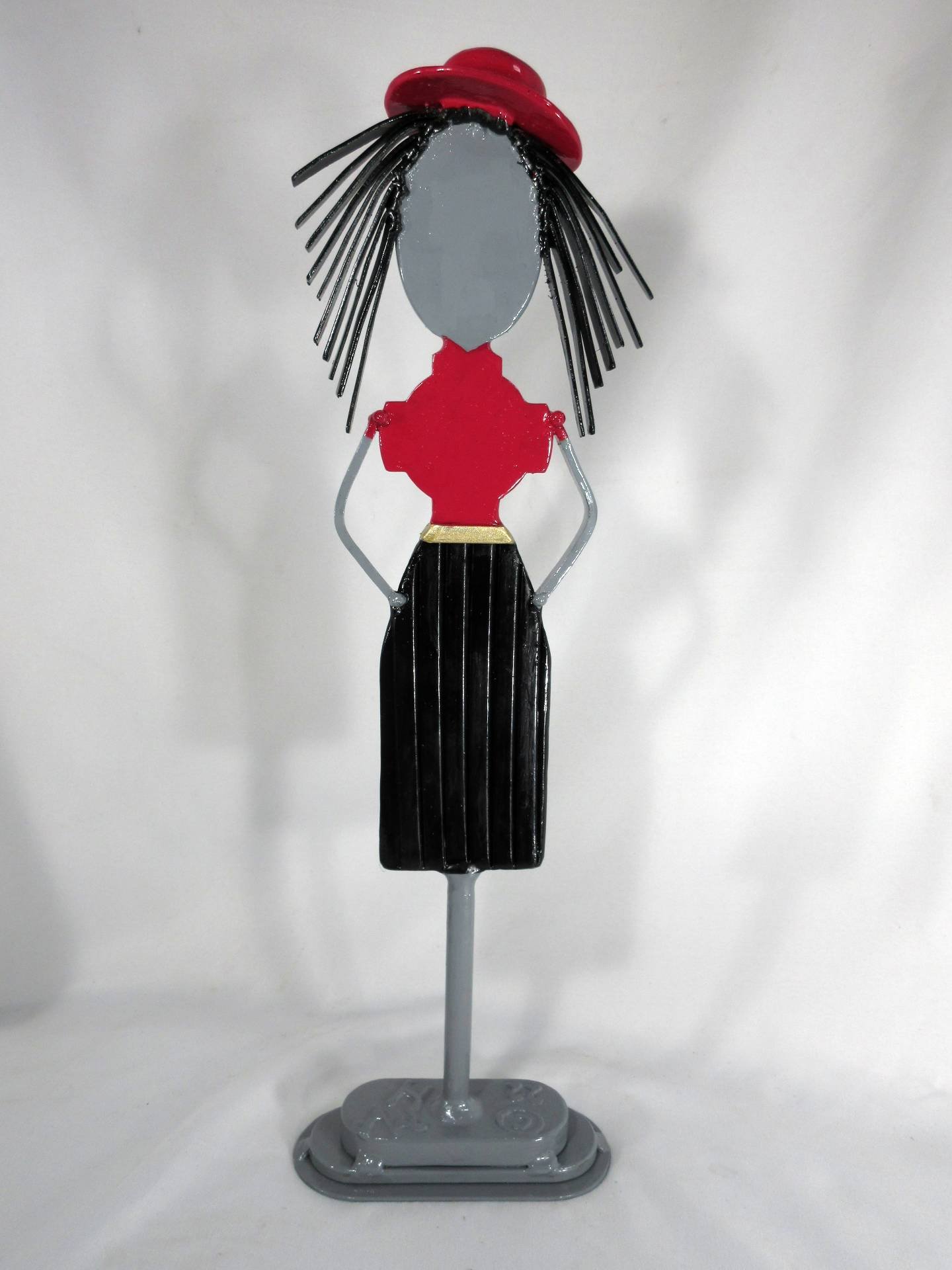 Stylized metal sculpture of a female figure with a red top, black skirt, and hat, standing upright with simplified facial features and linear hair elements. By Michael Herold.