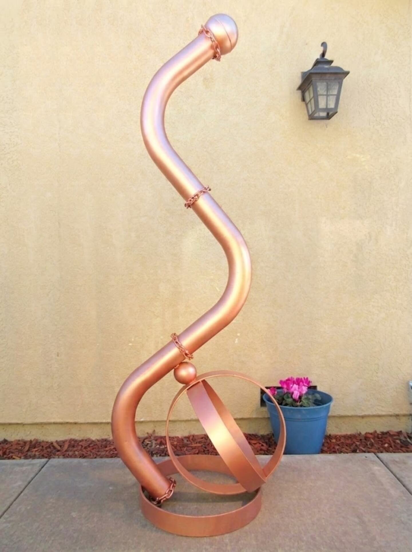 Curved copper-toned tubular sculpture rising in an S-shape, anchored to a circular base with a ring element and small sphere, featuring chain details along the structure. Michael Herold.