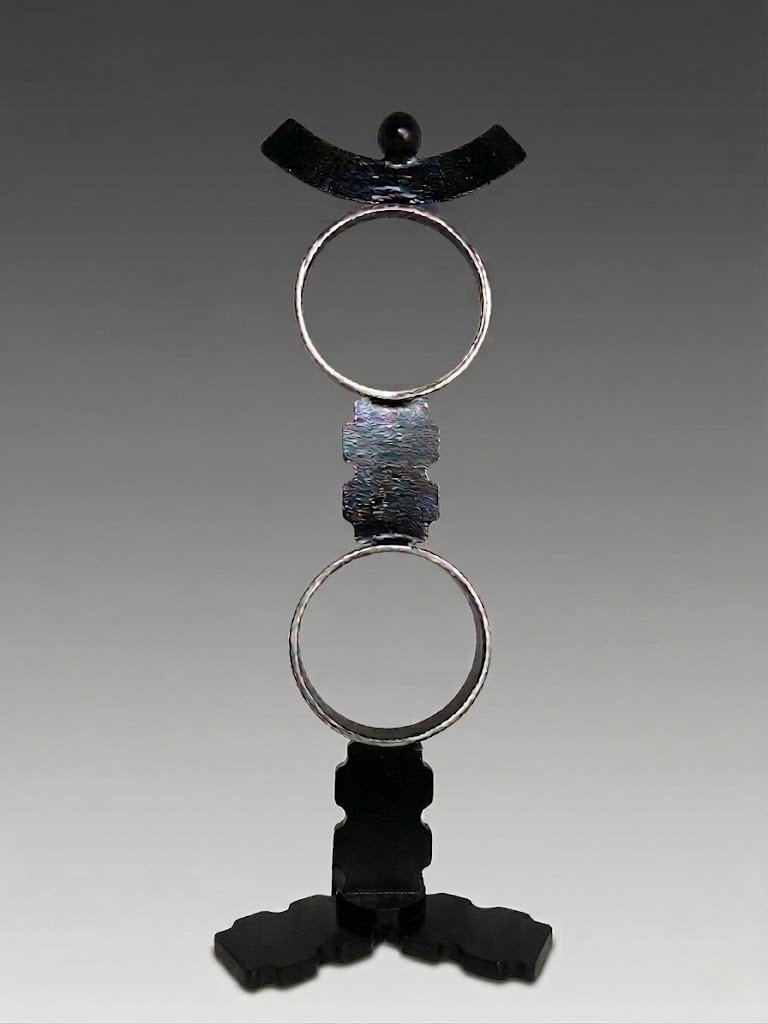 Vertical metal sculpture featuring two circular rings stacked along a central axis with textured connectors, topped by a curved element and small sphere on a base. By Michael Herold.