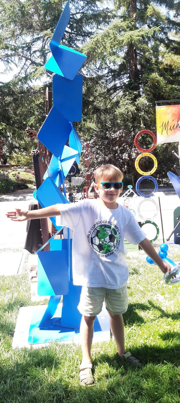 Young visitor posing in front of Michael Herold public sculpture at outdoor art festival, demonstrating playful engagement and emotional interaction with public art.