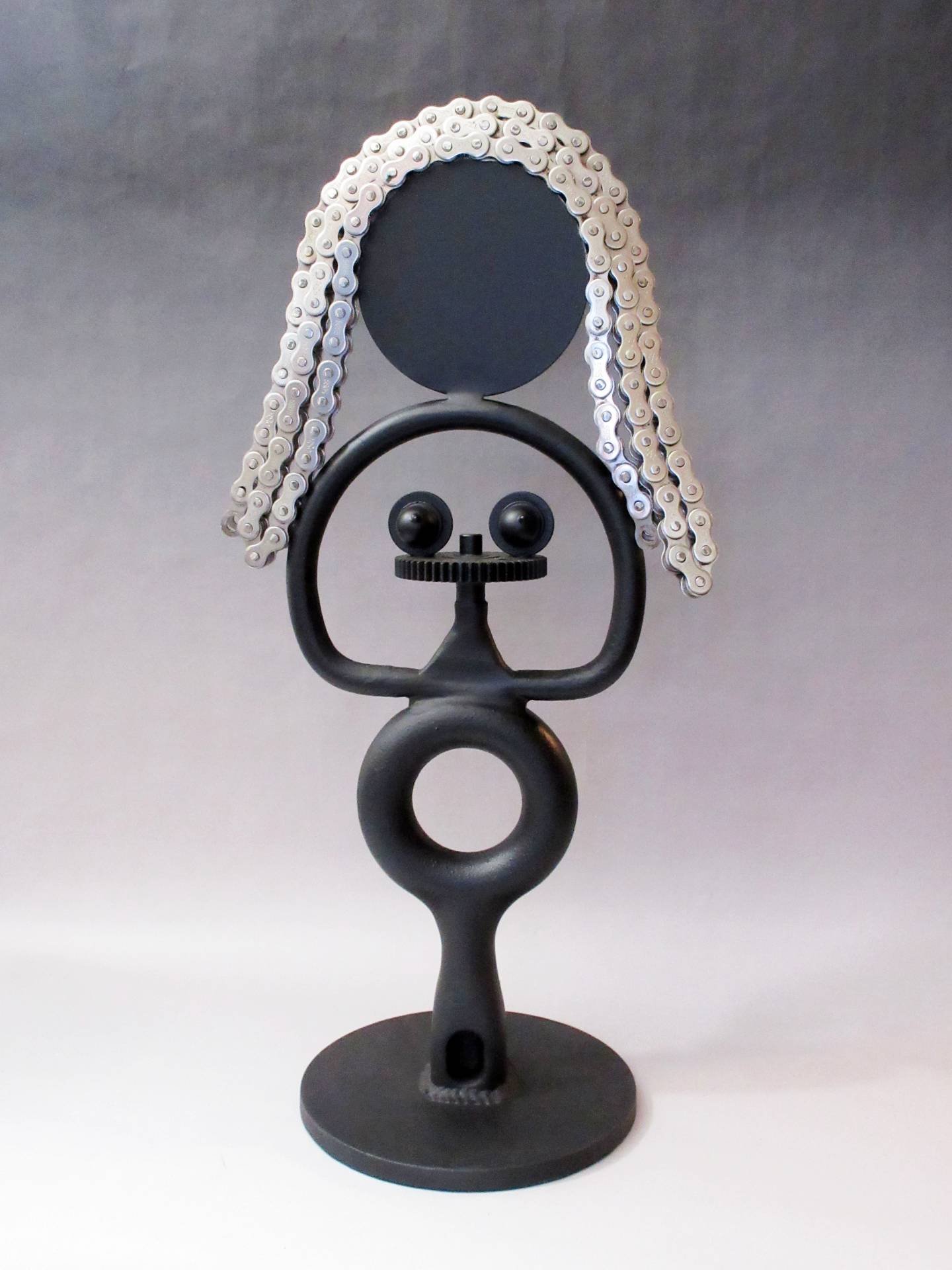 Abstract metal sculpture of a stylized human-like figure with circular forms, gear-like facial elements, and chain-like strands resembling hair. By Michael Herold.