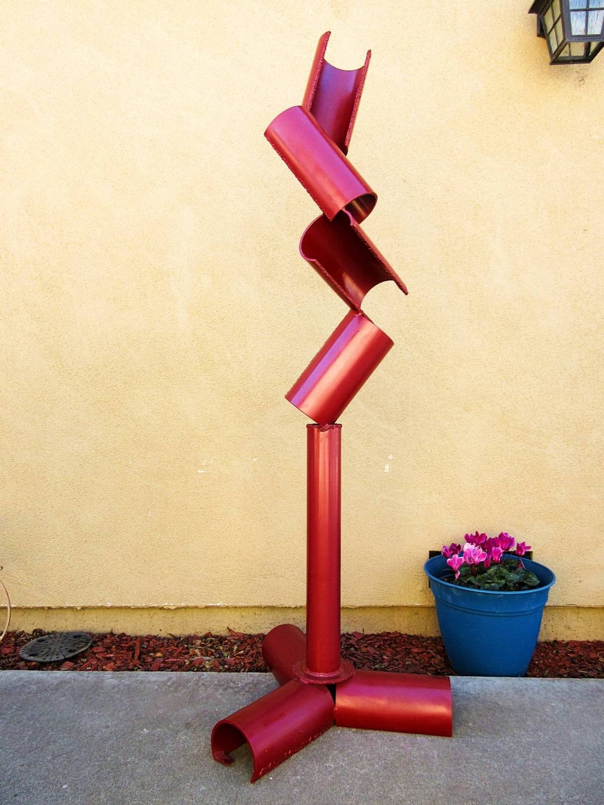 Red metal sculpture composed of curved cylindrical segments arranged in a tilted vertical structure on a three-part base.
