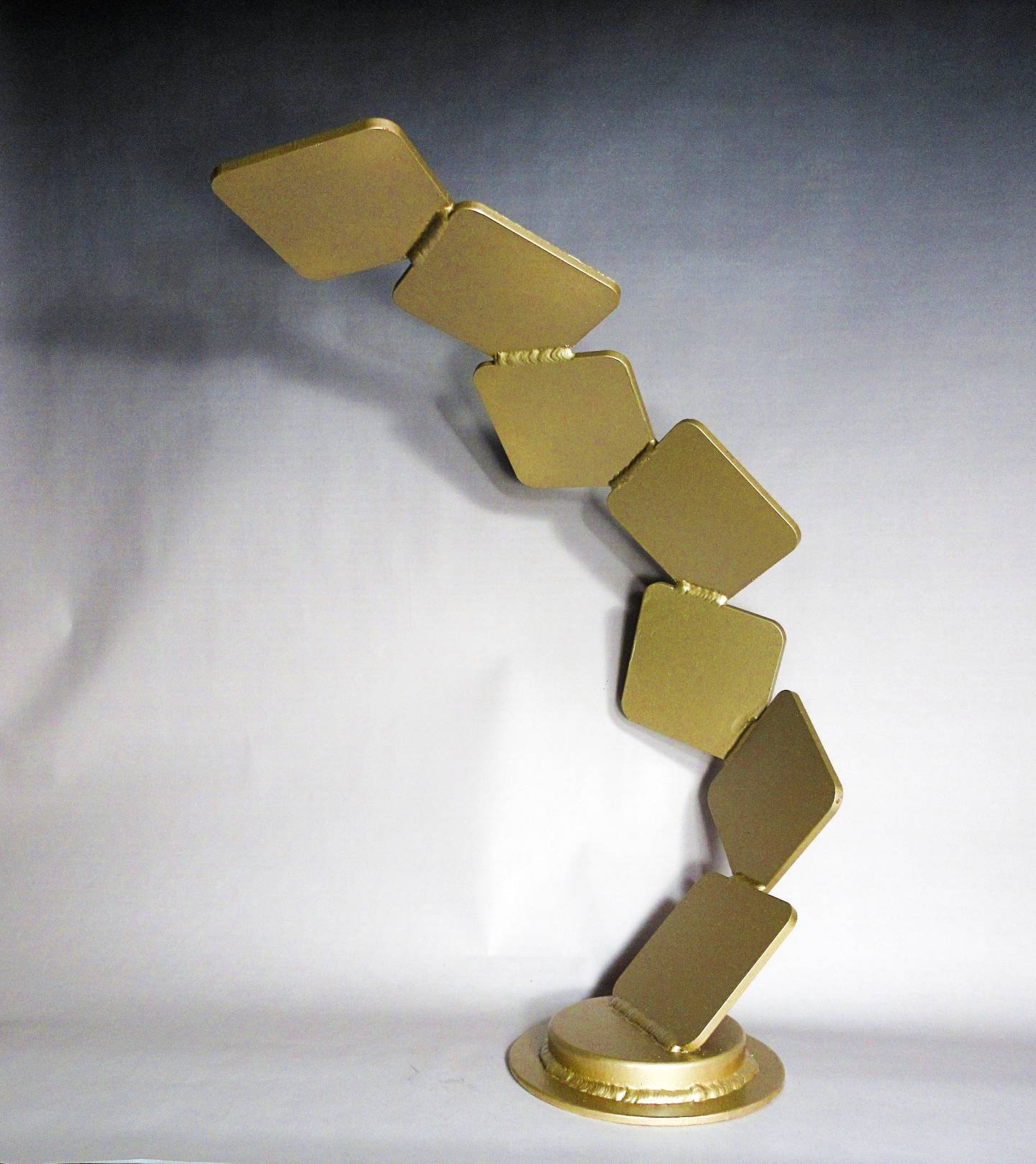 Gold metal sculpture composed of connected rectangular segments forming a curved, leaning structure on a circular base.