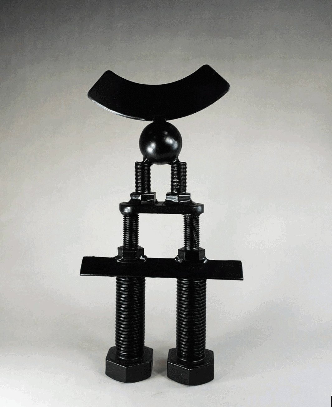 Black metal sculpture composed of stacked threaded rods, plates, and a central sphere, topped with a curved horizontal element, forming a symmetrical vertical structure. By Michael Herold.