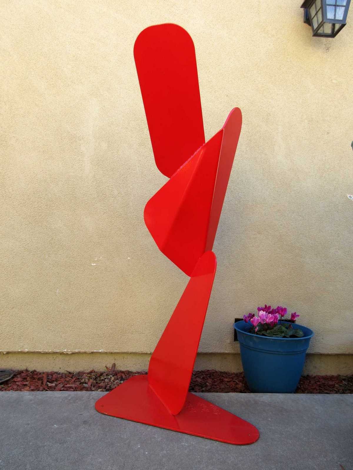 Red metal sculpture composed of angular, folded planes forming a vertical, forward-leaning structure on a flat base. By Michael Herold.