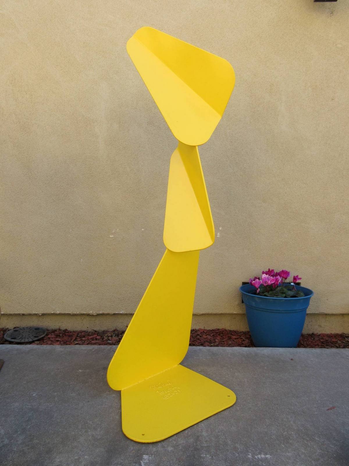 Yellow metal sculpture composed of angular, folded planes forming a vertical, forward-leaning structure on a flat base. By Michael Herold.